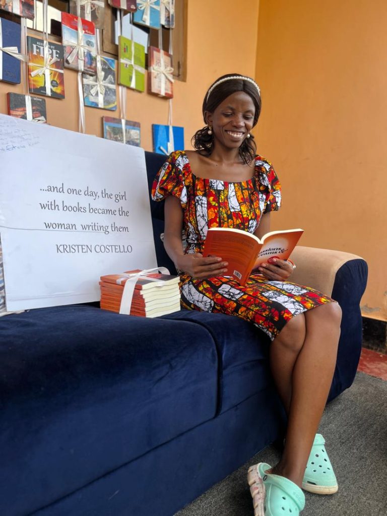 This is a photo of Winnie Naigaga taken on December 14, 2024, the day she launched her first book Passions and Dreams. In this photo, Winnie is seated on a dark blue couch holding a copy of Passions and Dreams with her head bent pretending to read the words she knows by heart. Her face is ardoned with a broad smile. Besides her is a pile of her books and a white cardboard with Kristen Costello's quote, "And the girl with books became the woman writing them."